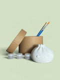 Paint Your Own Dumpling Kit with kraft box, paint pots and brushes on sage background